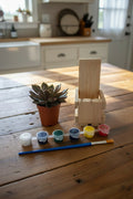 A miniature painting kit on a wooden tabletop, featuring a small wooden crate made from pallet wood, a small potted succulent, a paint palette with six colors, and a paintbrush.