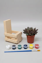A miniature wooden painting set displayed on a wooden tabletop against a light gray wall. The set includes a small wooden crate made from pallet wood, a potted succulent, five miniature paint pots in white, gray, green, yellow, and red, and a small blue-handled paintbrush.