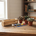 A wooden miniature painting kit displayed on a light wood table against a neutral wall, featuring a small wooden frame made from pallet wood, three potted succulent plants, a paintbrush, and small paint pots.