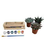 A miniature wooden planter set on a light wood table against a gray wall, featuring a small wooden crate made from pallet wood, a paint palette with five small pots in white, blue, green, yellow, and red, a paintbrush, and three small potted succulents.