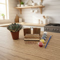 A miniature succulent plant in a small terracotta pot displayed alongside miniature building materials, including two wooden blocks made from reclaimed pallet wood, several pieces of reclaimed pallet wood, a small blue paintbrush, and tiny pots of colorful paint and a row of small nails, all arranged on a light wood-grained tabletop against a plain wall.