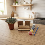 A miniature succulent plant in a small terracotta pot displayed alongside miniature building materials, including two wooden blocks made from reclaimed pallet wood, several pieces of reclaimed pallet wood, a small blue paintbrush, and tiny pots of colorful paint and a row of small nails, all arranged on a light wood-grained tabletop against a plain wall.