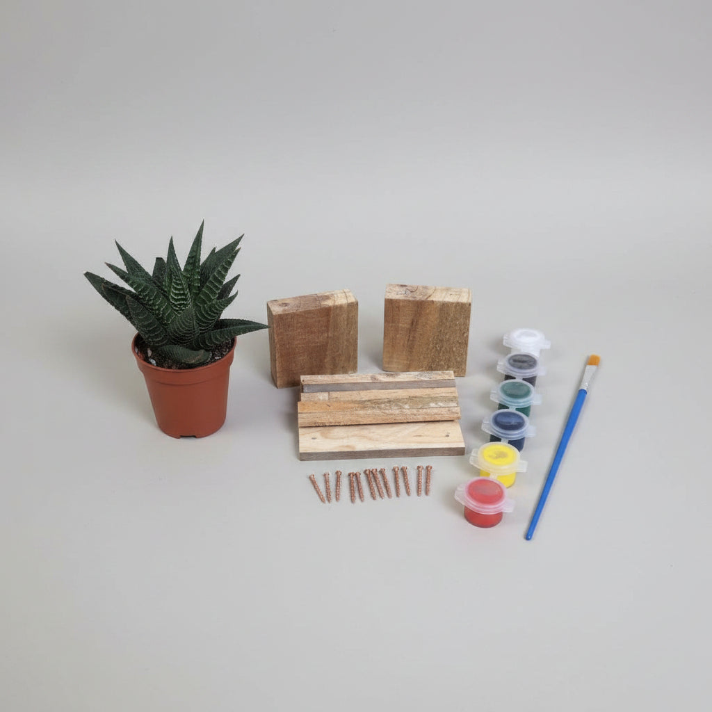 A miniature wooden block and nail kit displayed on a light wood-grained tabletop next to a small potted succulent. The kit includes various sizes of wooden blocks, copper-colored nails, small paint pots in multiple colors, and a blue-handled paintbrush.